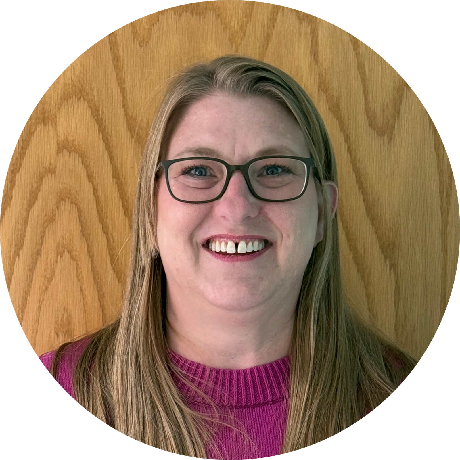 A woman with long, straight blonde hair and glasses smiles at the camera. She is wearing a magenta sweater and standing in front of a light wood-paneled background.