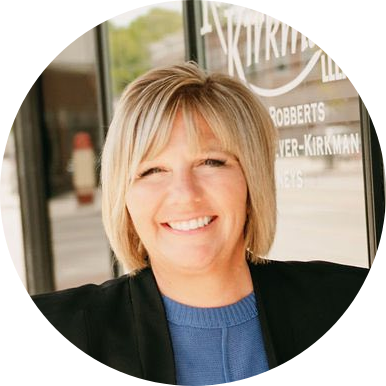 A woman with short blonde hair, wearing a blue top and black blazer, smiles while standing outside in front of a glass window with white text.