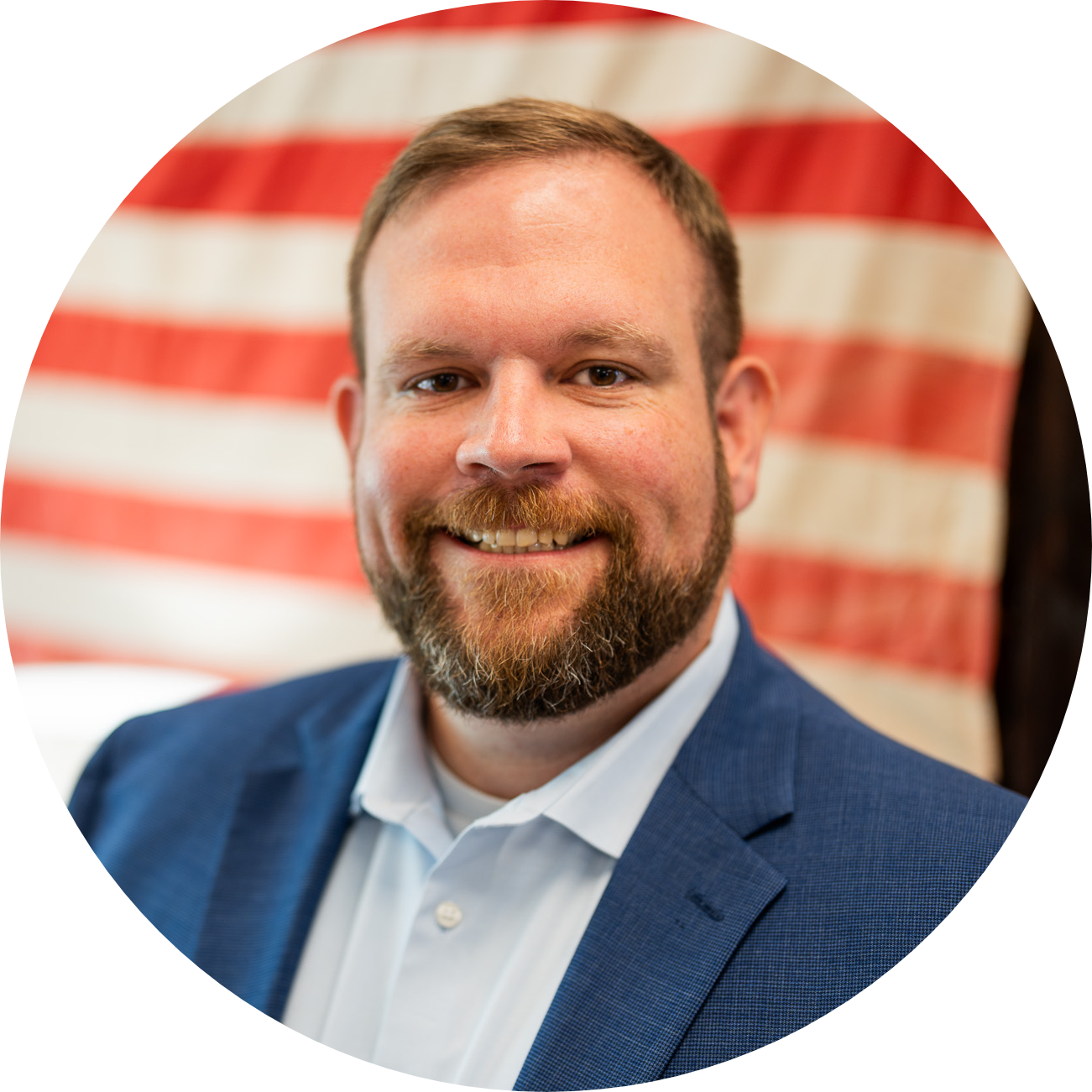 A man with a beard and short hair, wearing a blue suit jacket and light blue shirt, smiles at the camera. An American flag is blurred in the background.